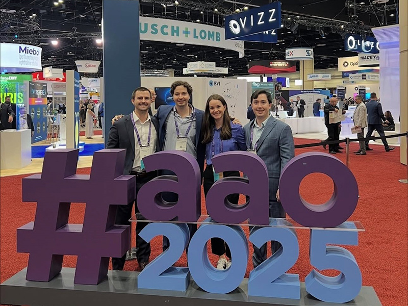 Four adults pose behind a large "#aao 2025" sign at a convention center with red carpet flooring. Exhibition booths for companies like "Bausch + Lomb" and "OVIZZ" are visible in the background, along with other attendees.